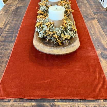 A rust-orange velvet table runner laid out on a dark wood dining table, styled with a wooden bowl centerpiece containing white candles and yellow-green floral accents.