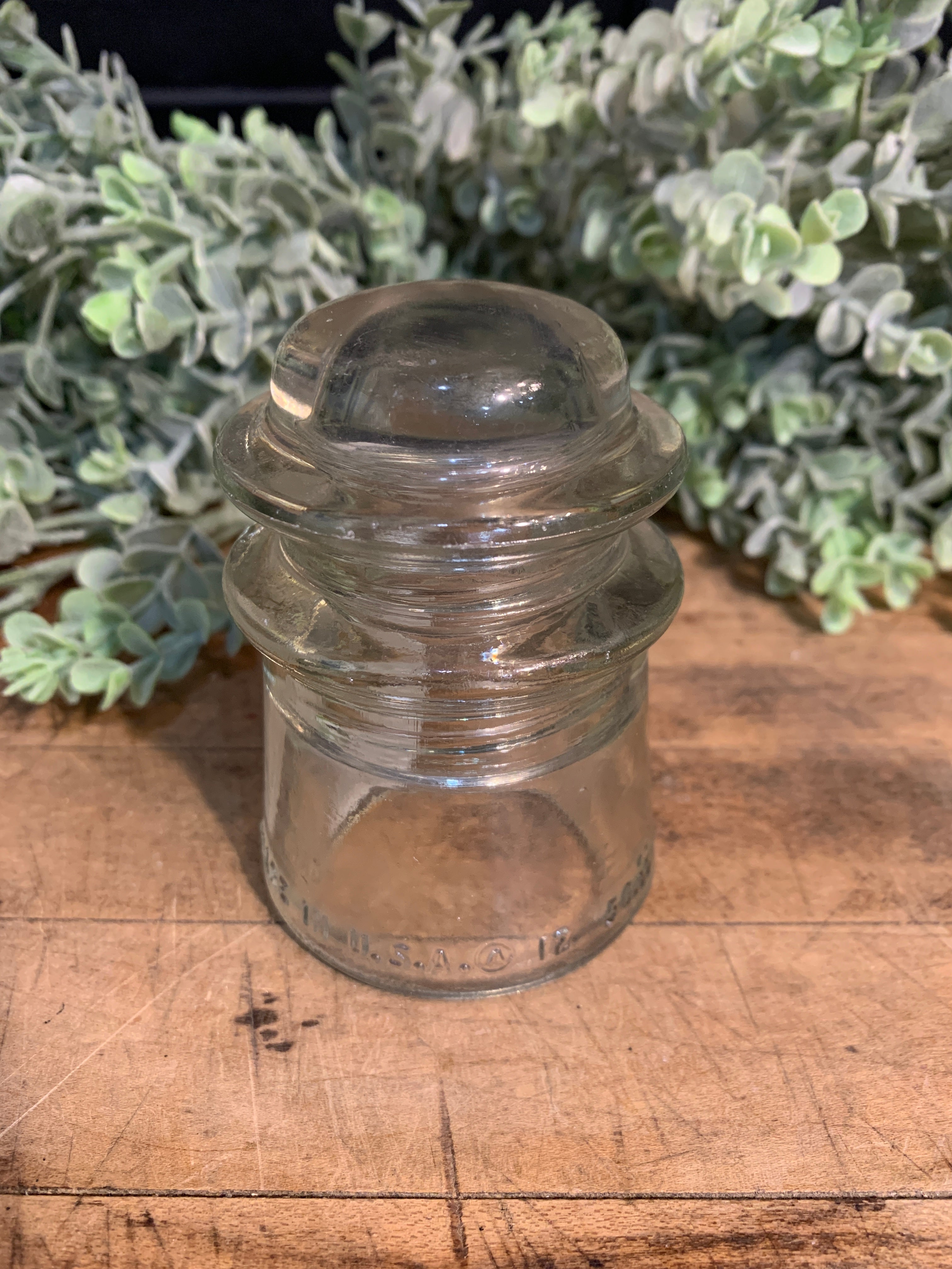 A vintage clear glass insulator displayed on a wooden surface with a plant in the background.