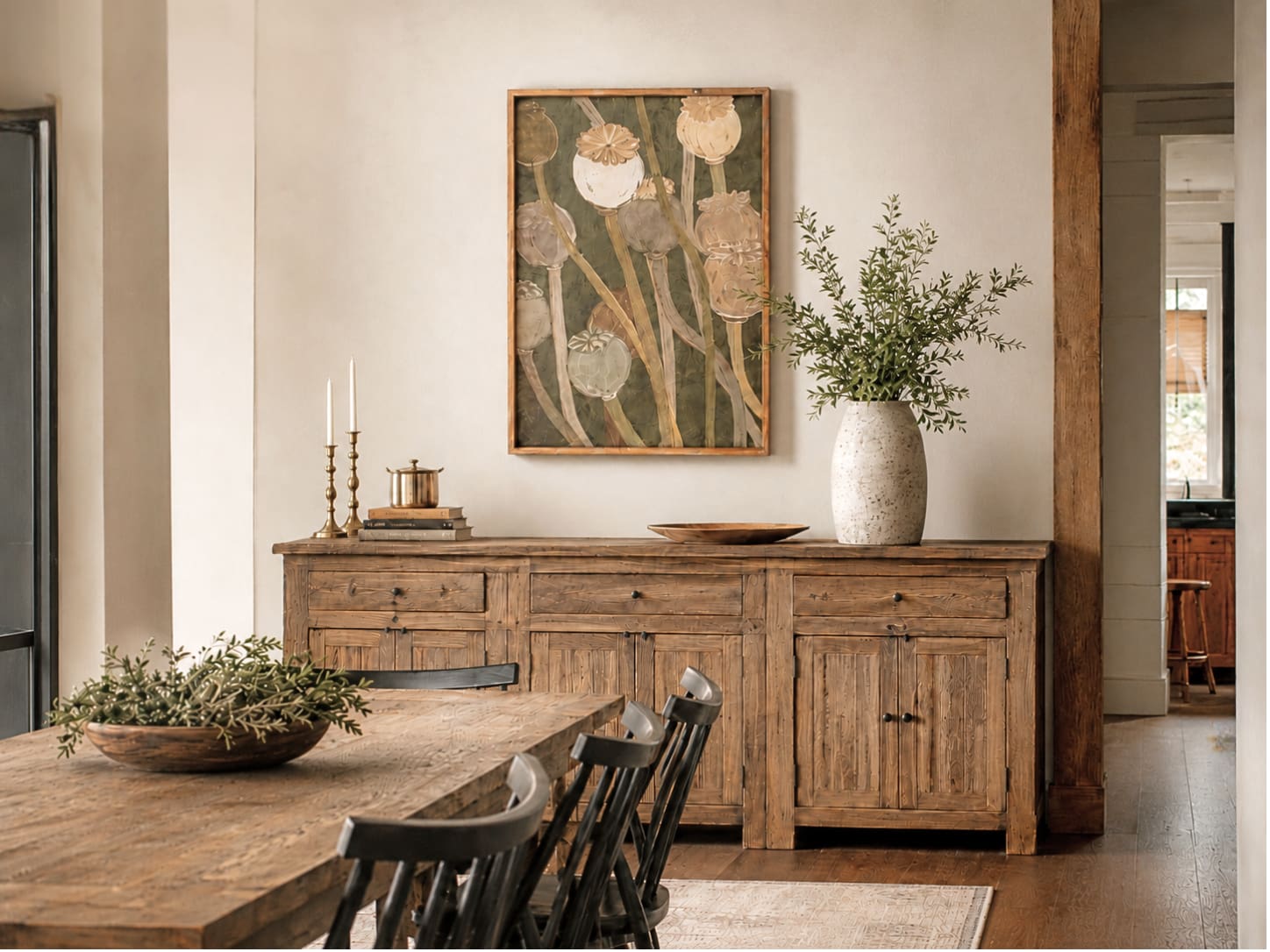 A rustic wooden sideboard in a warm dining room setting, styled with a framed botanical art print, a white vase with greenery, and candles.